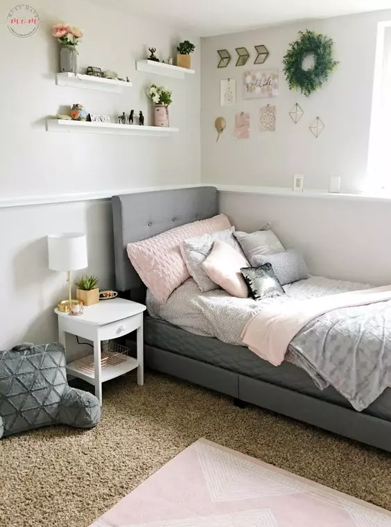 A modern teen bedroom with a grey upholstered bed and pastel bedding, layered rugs, wall mounted shelves and a greenery wreath