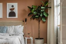 a boho bedroom with a terracotta accent wall, a bed with a woven bedding, a potted plant and a stool as a side table