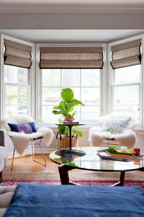 A bright boho sitting zone by the bay window   metal chais with pillows and faux fur, a potted plant and a window with Roman shades