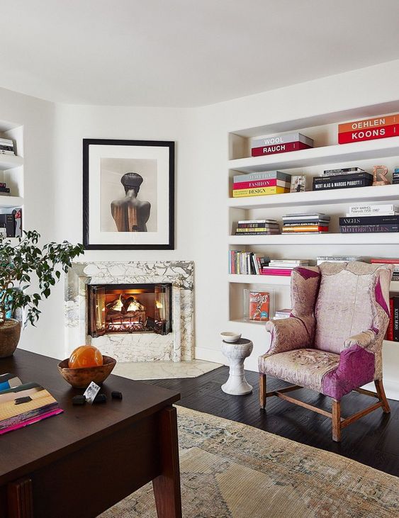 a chic and bold home office with a built-in fireplace with a marble surround, built-in bookshelves, a dark-stained desk and a colorful chair plus a neutral rug