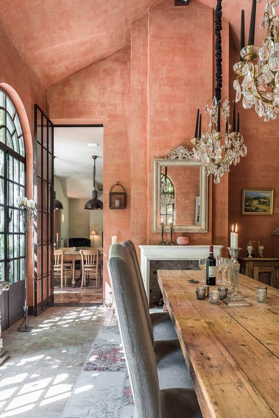 a jaw-dropping terracotta dining room with French windows, a fireplace, textural walls, a stained table and grey chairs plus crystal chandeliers