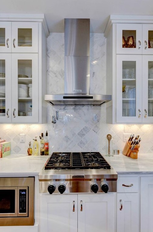 A modern white kitchen with upper glass cabinets, with a white marble arabesque tile backsplash and built in appliances is amazing
