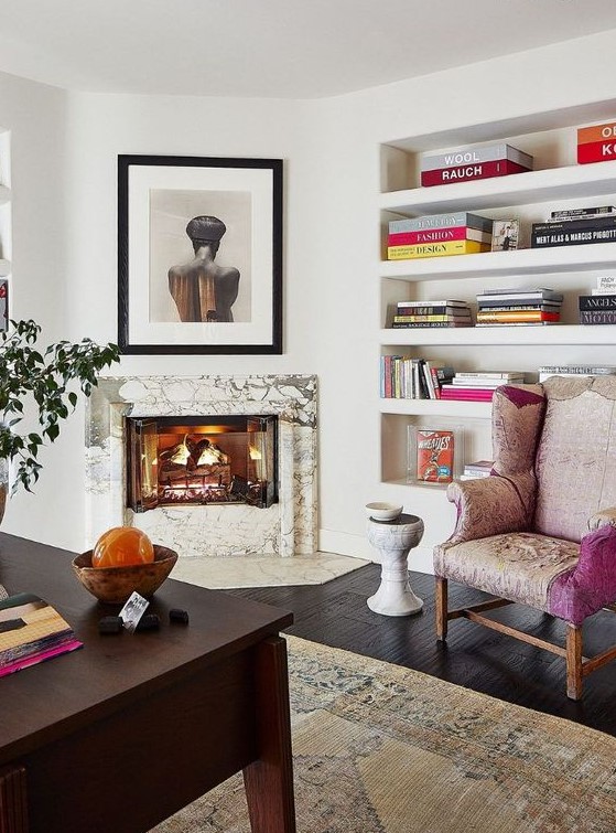 A refined home office with a marble clad fireplace, built in shelves, a dark stained desk, a pink chair and some art and bright books