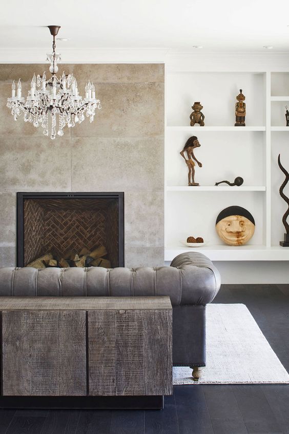 a refined lviing room with built-in shelves, a concrete fireplace, grey seating furniture, a wooden cabinter and a chic crystal chandelier