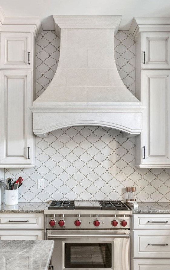 A refined vintage white kitchen with shaker style cabinets, a white arabesque tile backsplash and a vintage hood plus built in appliances