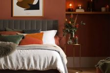 a soothing bedroom with terracotta walls, a grey upholstered bed with orange and white bedding, wall-mounted shelves and some lovely decor
