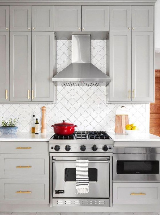 a stylish contemporary kitchen done in dove grey, with shaker cabinets, gold handles, a white arabesque tile backsplash and black grout