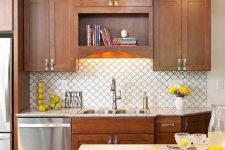 a stylish stained kitchen with a white arabesque tile backsplash, a neutral stone countertop, built-in lights and pendant lamps
