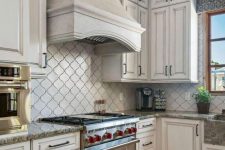 a vintage neutral kitchen with shaker cabinets, a grey stone countertop and a neutral arabesque tile backsplash, a large hood