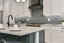 a vintage white kitchen with shaker cabinets, a grey kitchen island, white stone countertops, black fixtures and appliances and a grey arabesque tile backsplash