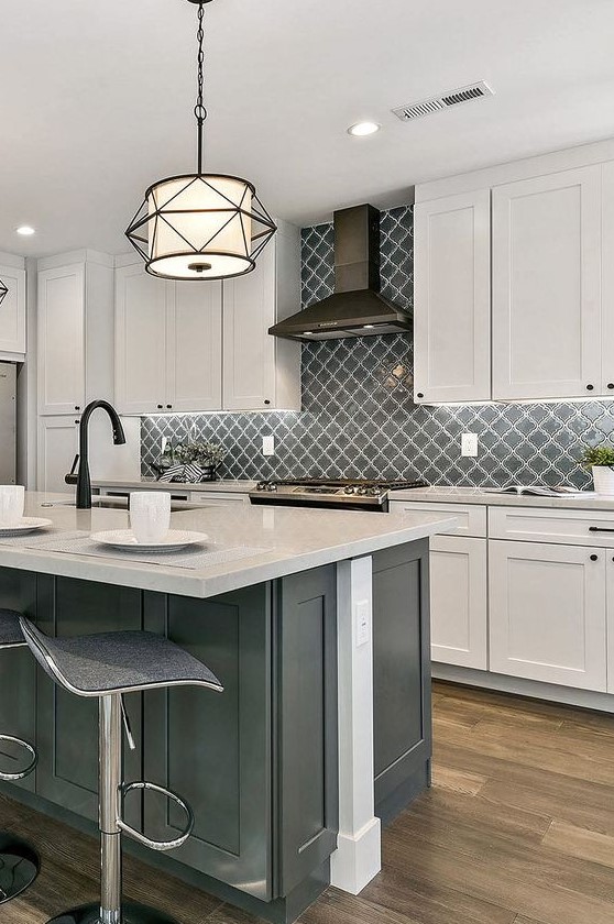 a vintage white kitchen with shaker cabinets, a grey kitchen island, white stone countertops, black fixtures and appliances and a grey arabesque tile backsplash