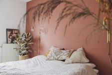 a welcoming bedroom with a terracotta accent wall, a bed with neutral bedding, gold sconces and potted plants and grasses