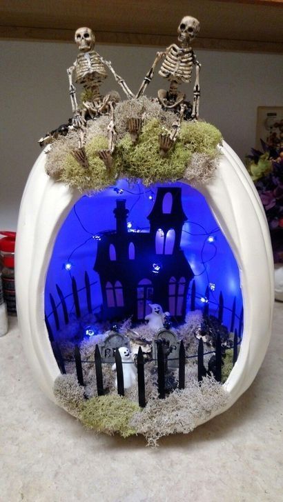 a white Halloween diorama with a blue inside, a black house, lights, a graveyard with ghosts and a couple of skeletons on top