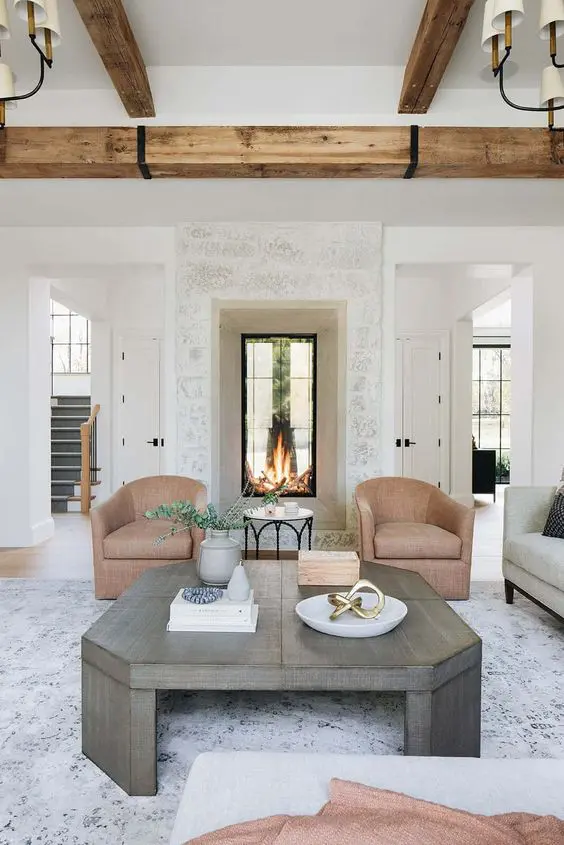 A beautiful neutral living room with a built in double sided fireplace, beige chairs, a grey coffee table and wooden beams