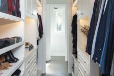 a narrow white walk-in closet with shelves, open storage compartments, railings, lights and lit up shelves