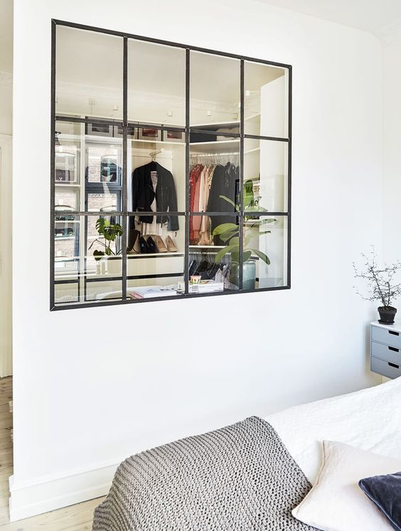 an airy Scandinavian bedroom is connected to the walk-in closet with a large window with black frames and it floods both spaces with natural light