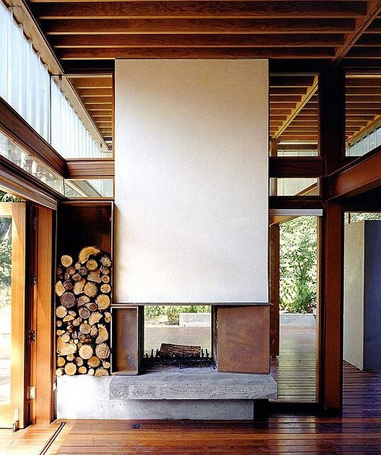 an ultra-modern double-sided fireplace with a white panel over it and metal parts plus firewood stored next to it