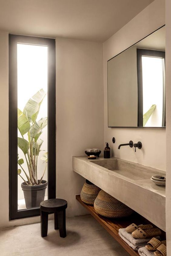 A concrete Japandi bathroom with a large mirror, a built in concrete vanity and a shelf, a vertical window plus a black stool