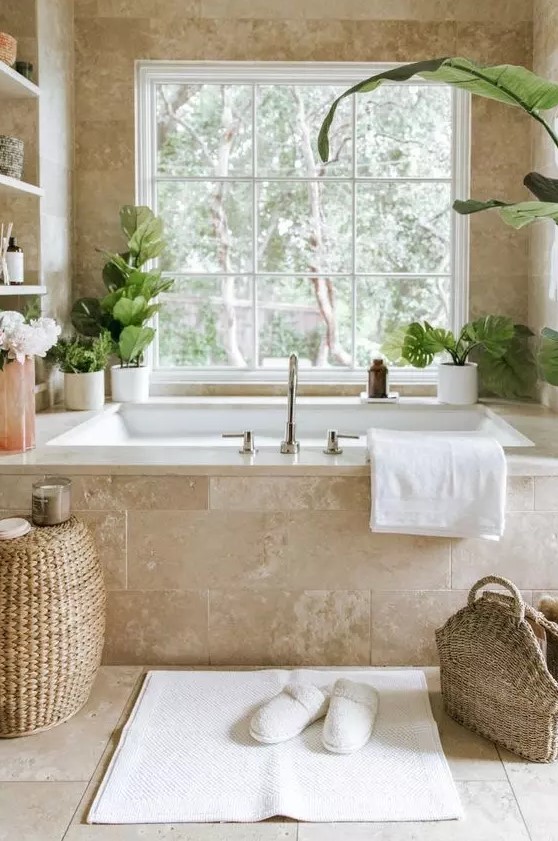 A gorgeous neutral spa bathroom clad with limestone tiles, with a tub clad with them, built in shelves and potted greenery, a woven pouf and a bag