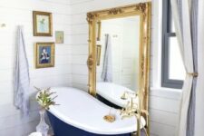 37 a refined bathroom with white shiplap, a navy clawfoot bathtub, a floor mirror in a gold frame, a crystal chandelier and artworks