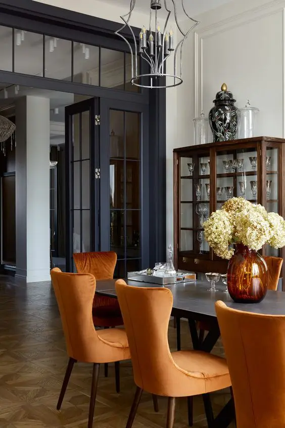 A refined dining room with a dark stained glass cabinet, a black table, orange chairs, large black French doors plus a matching transom window