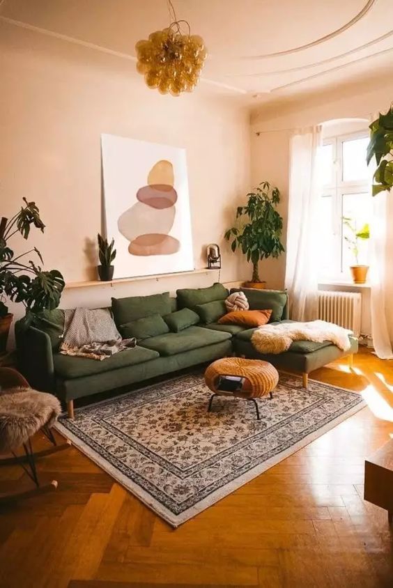 A muted and warm colored living room with tan walls and a ceiling, a green sectional, a woven pouf, a printed rug and potted plants