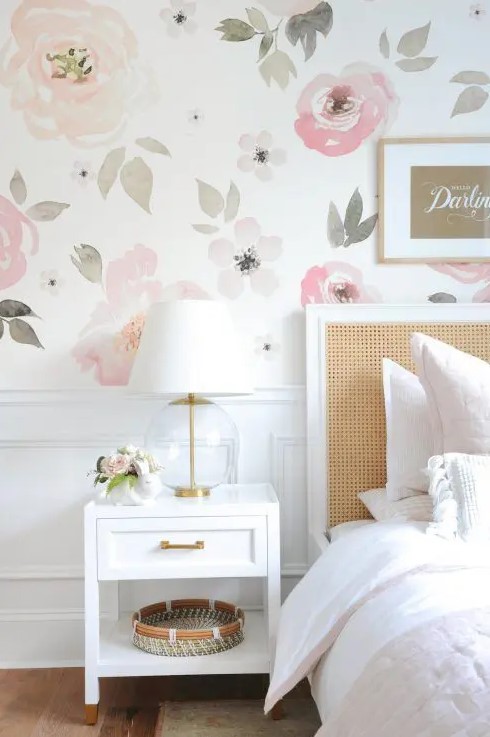 a lovely bedroom with a watercolor floral wall, a bed with a rattan headboard, white nightstands, gold touches