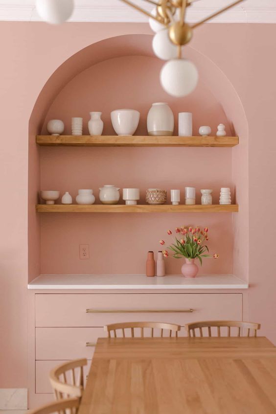 a pink dining room with an arched niche with stained shelves used to display vases and a built-in cabinet with drawers