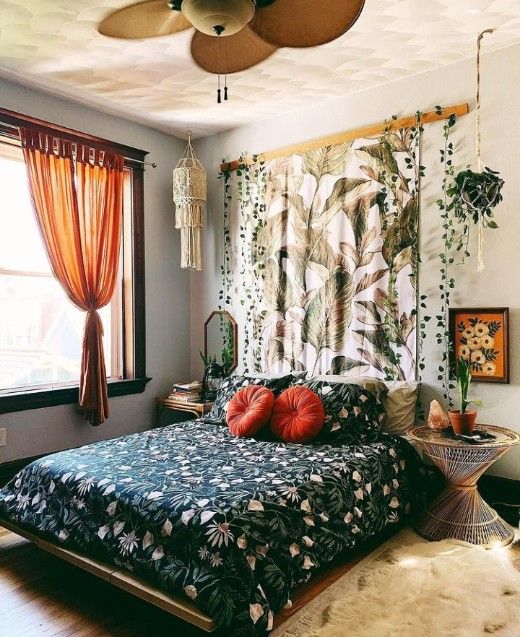 a boho bedroom with a bed with dark floral bedding, rattan nightstands, a botanical hanging, orange curtains and potted greenery