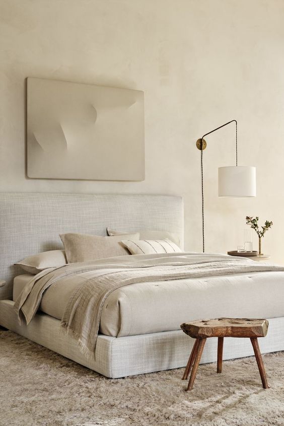 a neutral textural bedroom with limewashed walls, an upholstered bed, textural bedding, a catchy artwork and a wooden stool
