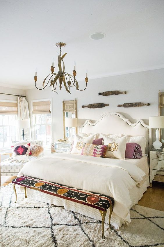 a neutral bedroom with an upholstered bed, printed pillows, a printed bench, a chair with printed pillows and a printed rug