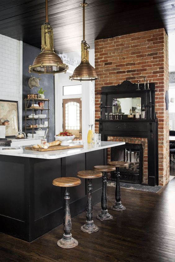 An industrial kitchen with a built in fireplace, a black kitchen island with a white countertop, tall vintage stools and brass pendant lamps