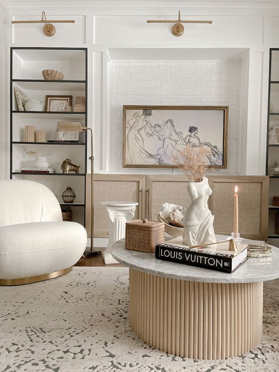 a neutral living room with built-in niche shelves, a rattan console, a creamy chair, a marble coffee table with a fluted base