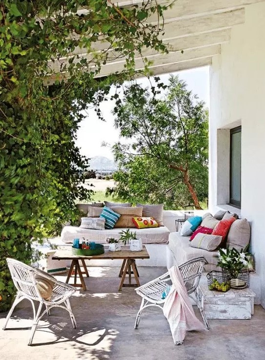 A lively Mediterranean outdoor space with a white built in sofa with neutral upholstery and bright pillows, a wooden trestle table and wicker chairs, greenery weaving around the pillars
