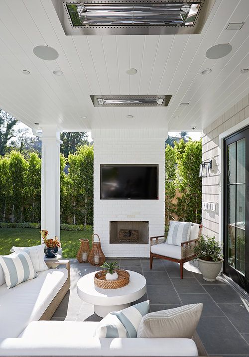 A neutral outdoor living room with a built in fireplace, a neutral sofa and chairs, a white round coffee table and woven lanterns