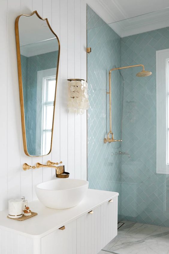 a beautiful neutral and light blue bathroom, with a white floating vanity, brass fixtures and a cool mirror