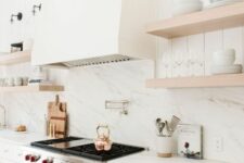 a creamy and airy kitchen with shaker cabinets, a white quartz backsplash and countertops, wooden shelves and a large hood