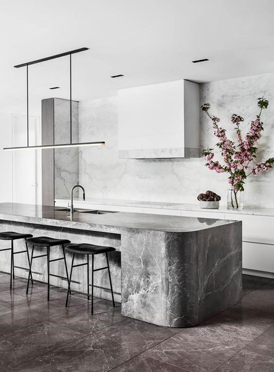 a minimalist white kitchen with a grey curved kitchen island made of a marble slab and a white marble backsplash and countertops