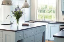 a modern country light blue  kitchen with dark countertops, pendant lamps and large windows plus a lovely hardwood floor is chic