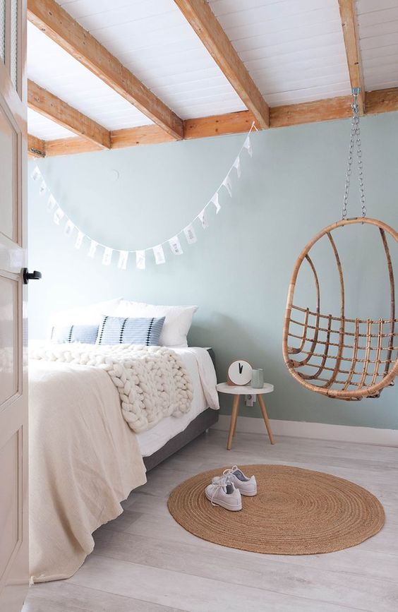 a pastel blue bedroom with a grey bed and neutral bedding, a rattan pendant chair, a small stool as a nightstand and a woven rug