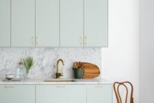 a refined light green kitchen with geo facades, a white marble backsplash and countertops plus gold fixtures