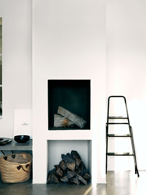 a white minimalist fireplace with firewood, a basket, a ladder and some decor around for a minimalist space