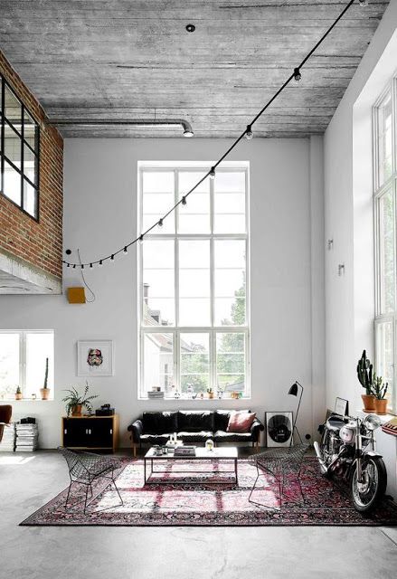 A boho double height living room with a wooden ceiling, double height windows, a black sofa, a coffee table, metal chairs, a bright rug and a bike