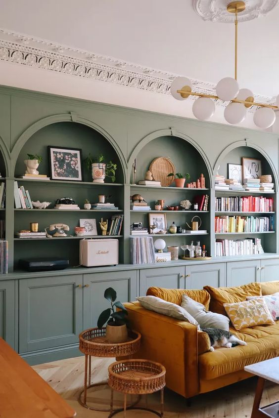 A chic living room with a sage green accent wall with built in niches and storage units, a mustard sofa, rattan side tables and lovely colorful niche decor