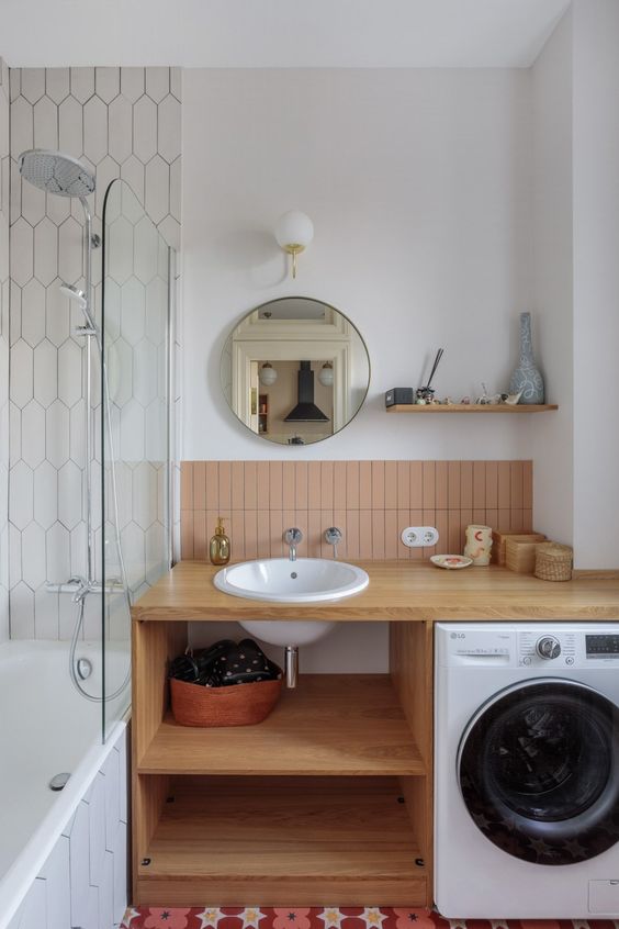 A mid century modern bathroom with a bathtub, an open timber vanity with a washing machine, dusty pink tiles and a round mirror