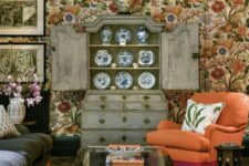 04 a maximalist cottagecore living room with bright floral wallpaper, a dark sofa and an orange chair, a black coffee table, a shabby chic buffet