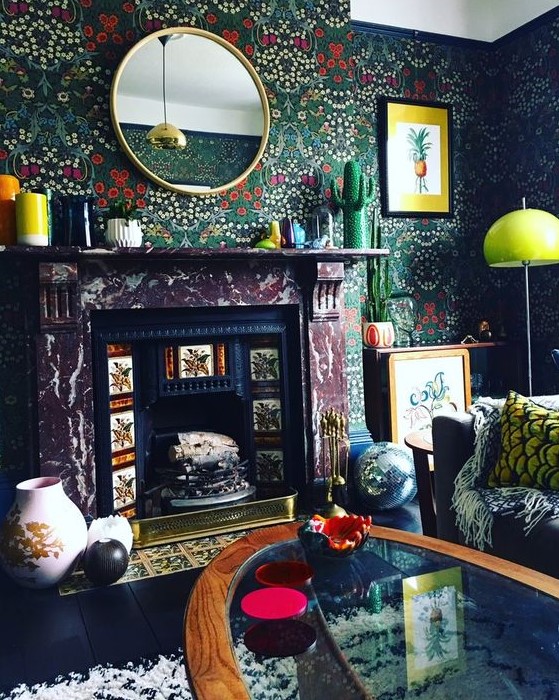 A moody living room with dark floral wallpaper, a non working fireplace, a black sofa and bold pillows, a glass coffee table