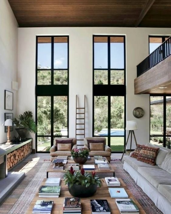 A neutral contemporary living room with double height windows, neutral seating furniture, large coffee tables, a fireplace and a firewood storage unit