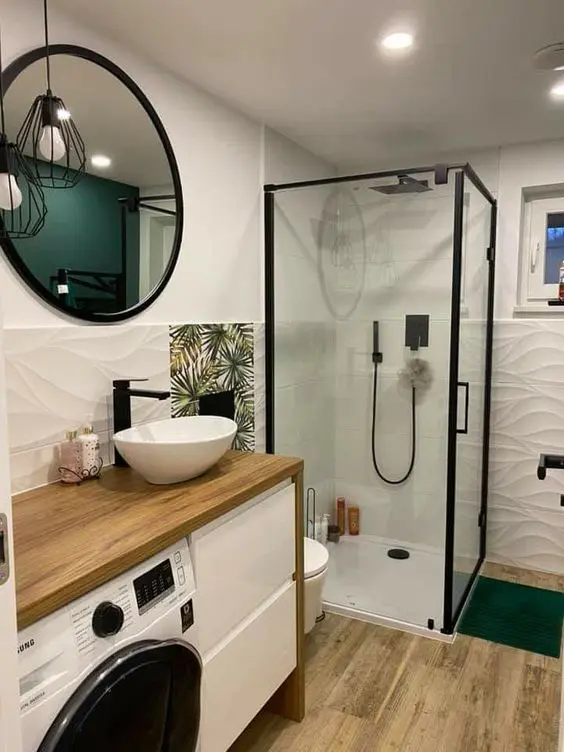 A small modern bathroom with a vanity and a built in washing machine, a glass enclosed shower, white textural tiles and a wooden floor