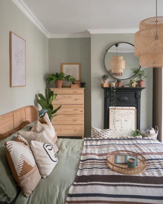 a sage green bedroom with stained furniture, printed bedding, a non-working fireplace, a woven pendant lamp and potted greenery
